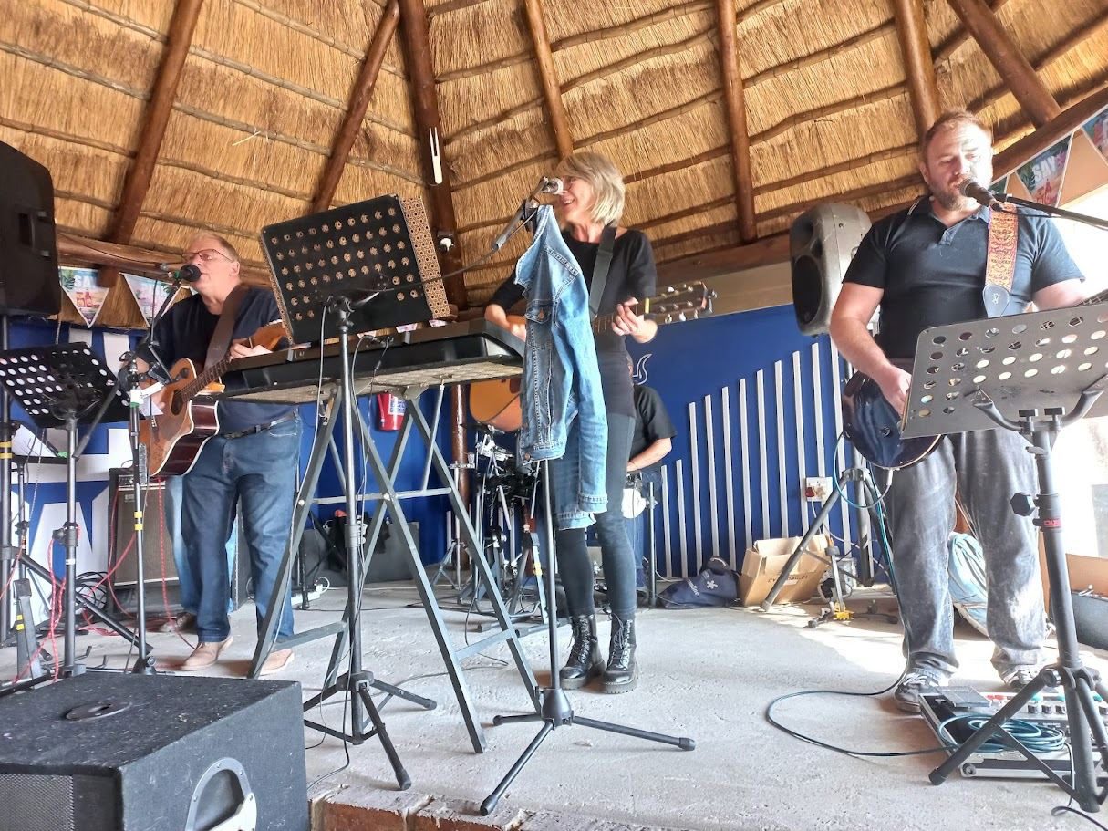 A four-piece band playing under a thatched roof, Warren on the right behind a music stand of chord sheets, another guitarist on the left
