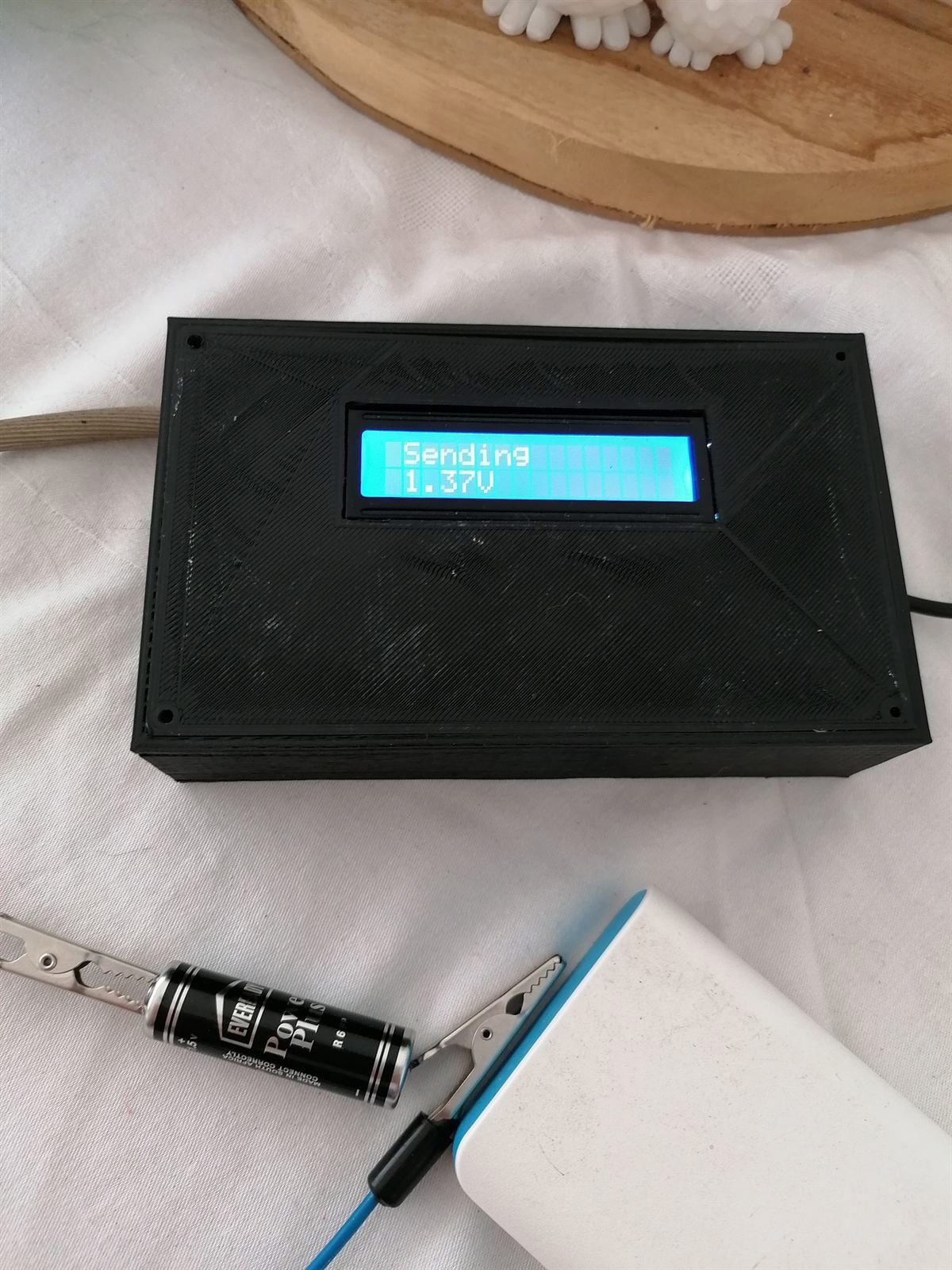 The finished monitor in its 3D-printed box, LCD reading Sending 1.37V, croc-clipped to a single AA battery