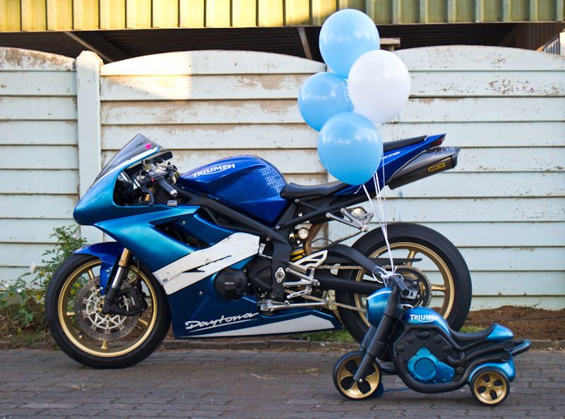A full-size blue Triumph Daytona parked next to a tiny matching blue and gold plastic ride-on bike, with balloons