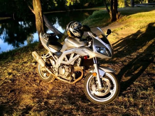 A silver Suzuki SV650S parked on the grass beside a river in late afternoon light, with a helmet resting on the seat