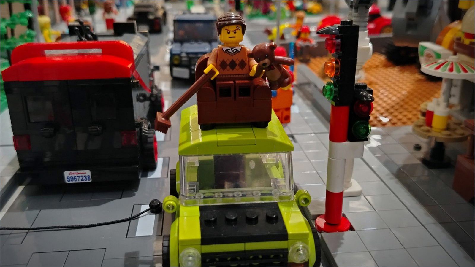 A scowling LEGO minifig in a brown armchair strapped to the roof of a lime-green Mini, stuck at a red traffic light in a LEGO city