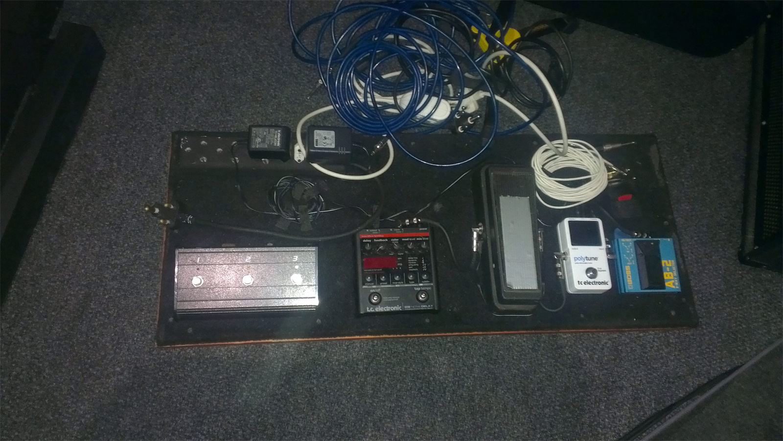 A guitar pedal board on a dark stage floor: five pedals chained together, with a tangled pile of blue and white cables heaped across the top of it