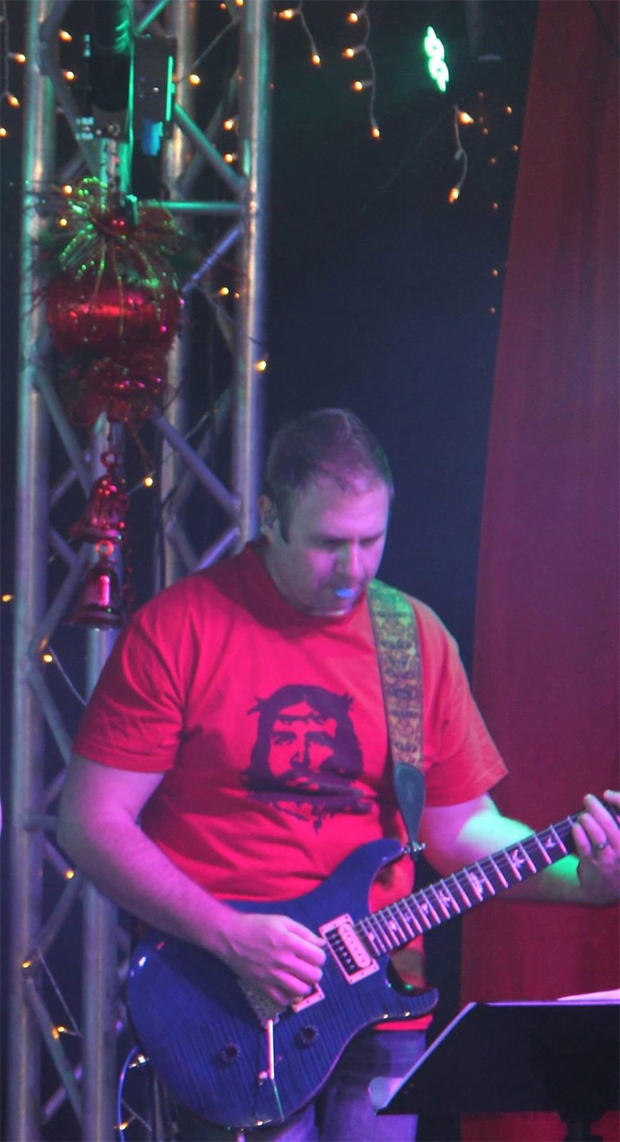 Warren on stage playing a blue electric guitar, head down, with lighting truss and cabling directly above him